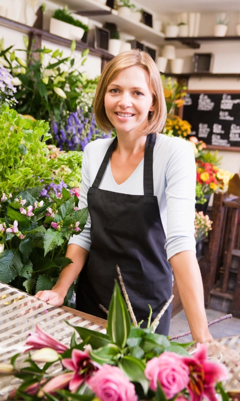 Lächelnde Floristin in Schürze steht hinter dem Verkaufstisch, umgeben von bunten Blumen.