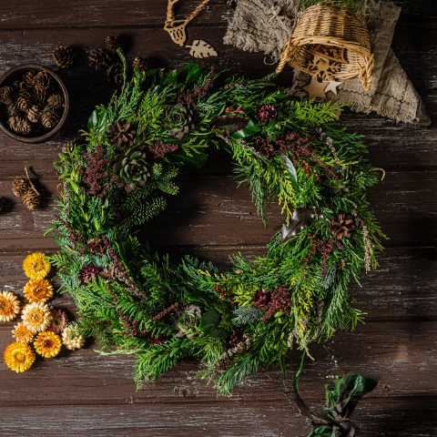 Dekorativer Adventskranz mit Tannengrün, Zapfen und Trockenblumen, rustikal auf Holzplatte arrangiert.