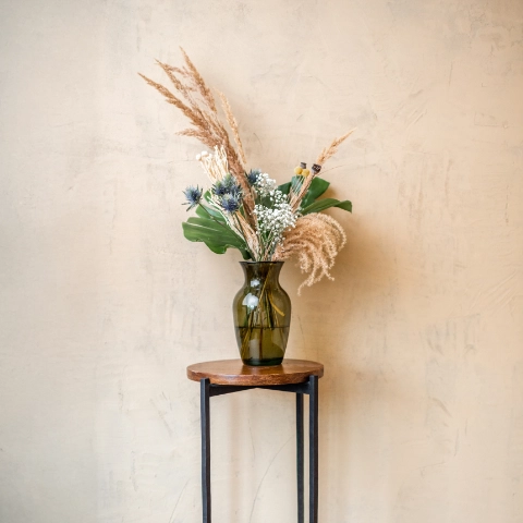 Dekorativer Trockenblumenstrauß in grünlicher Glasvase, auf schlichtem Holzhocker vor beigem Hintergrund arrangiert.