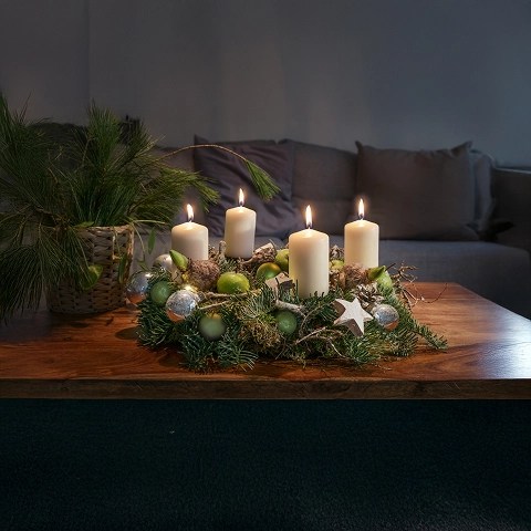 Weihnachtlicher Tischschmuck mit vier brennenden Kerzen, Tannenzweigen und silbernen Kugeln, in Wohnzimmeratmosphäre.